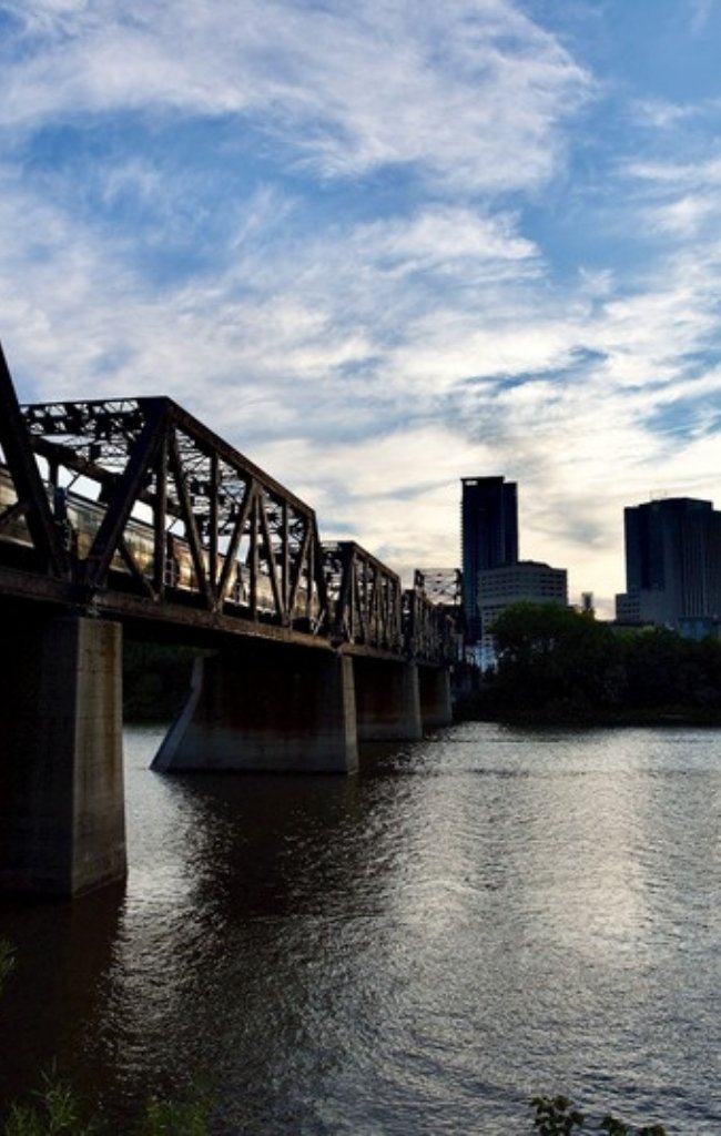 A bridge over the Red River with the Winnipeg skyline at sunset