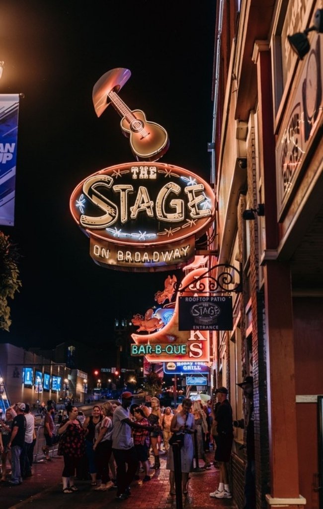 A neon guitar sign glowing above a crowded Broadway sidewalk