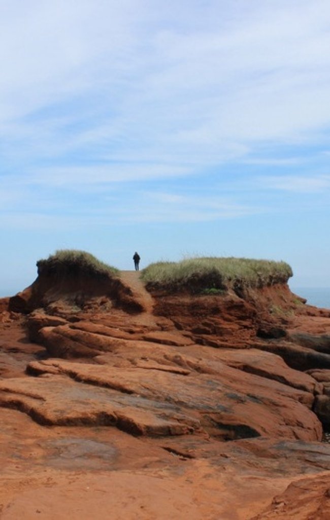 A person standing on red rock cliffs above a calm sea in Prince Edward Island