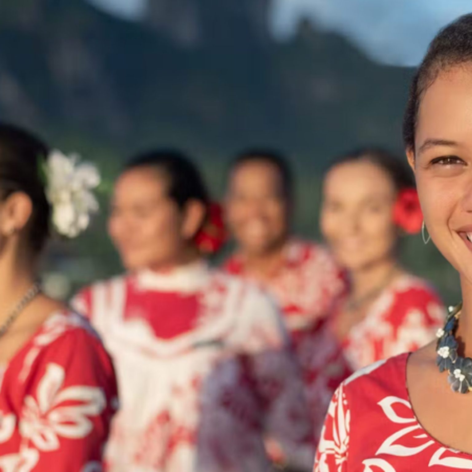 A Tahitian hostess in a red floral dress smiling at sunset