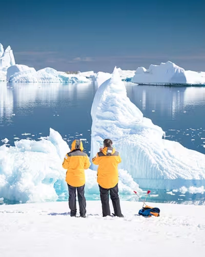 Two travelers in yellow parkas looking at icebergs in Antarctica