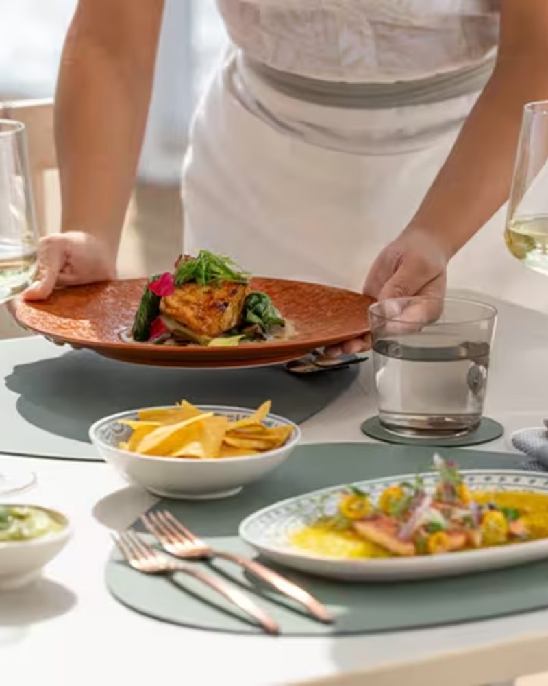 A server presenting a plated dish at a table set for lunch