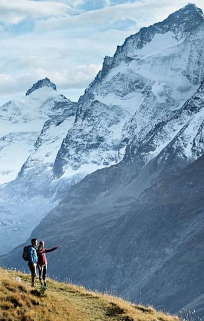 Two hikers on a grassy ridge facing snow-covered Alpine peaks
