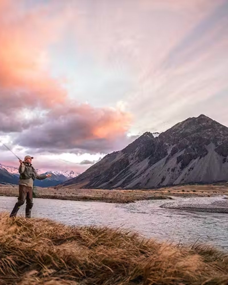 Angler fly fishing a braided river below New Zealand mountains at sunset